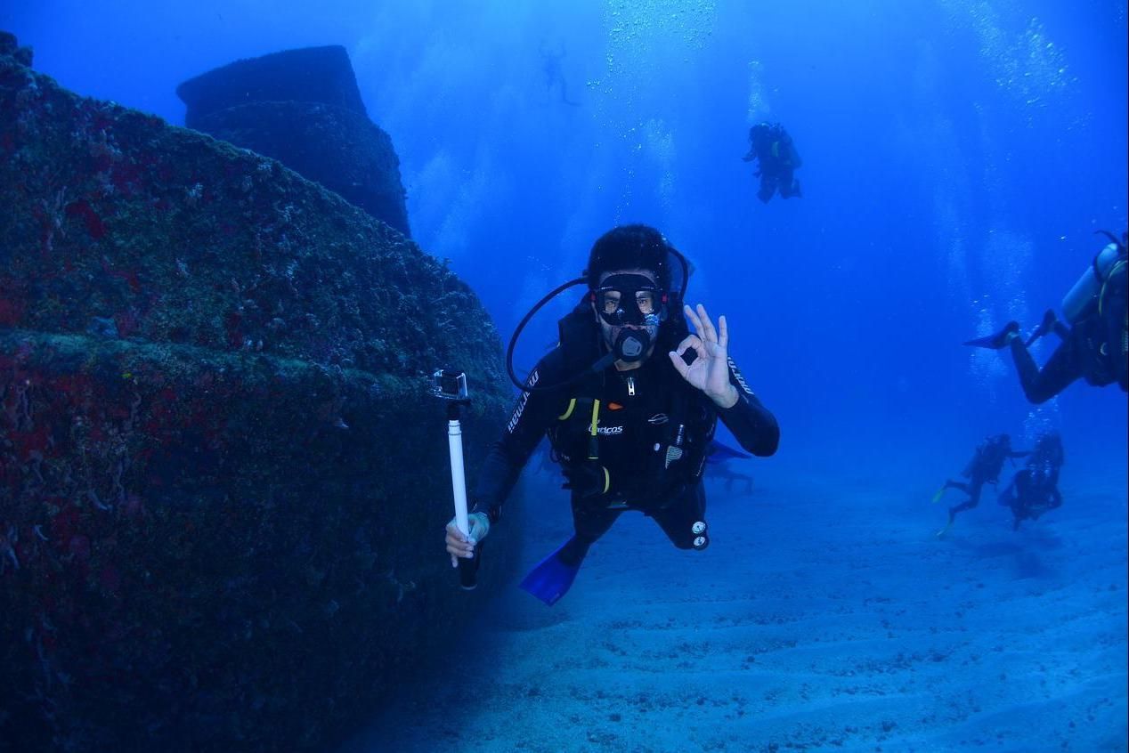 Scuba diver underwater beside a shipwreck, holding a camera and making an OK sign, with other divers nearby