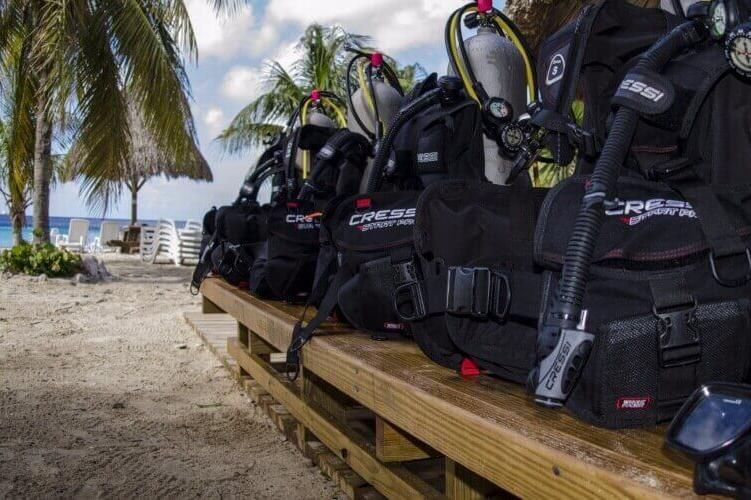 Black scuba gear bags and tanks on a wooden dock by palm trees and turquoise water