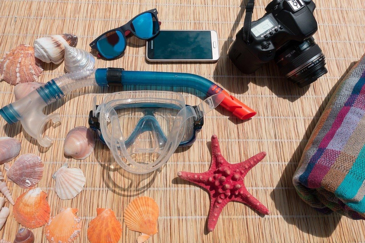 Snorkel mask, camera, sunglasses, phone, shells, and starfish on a sandy beach background