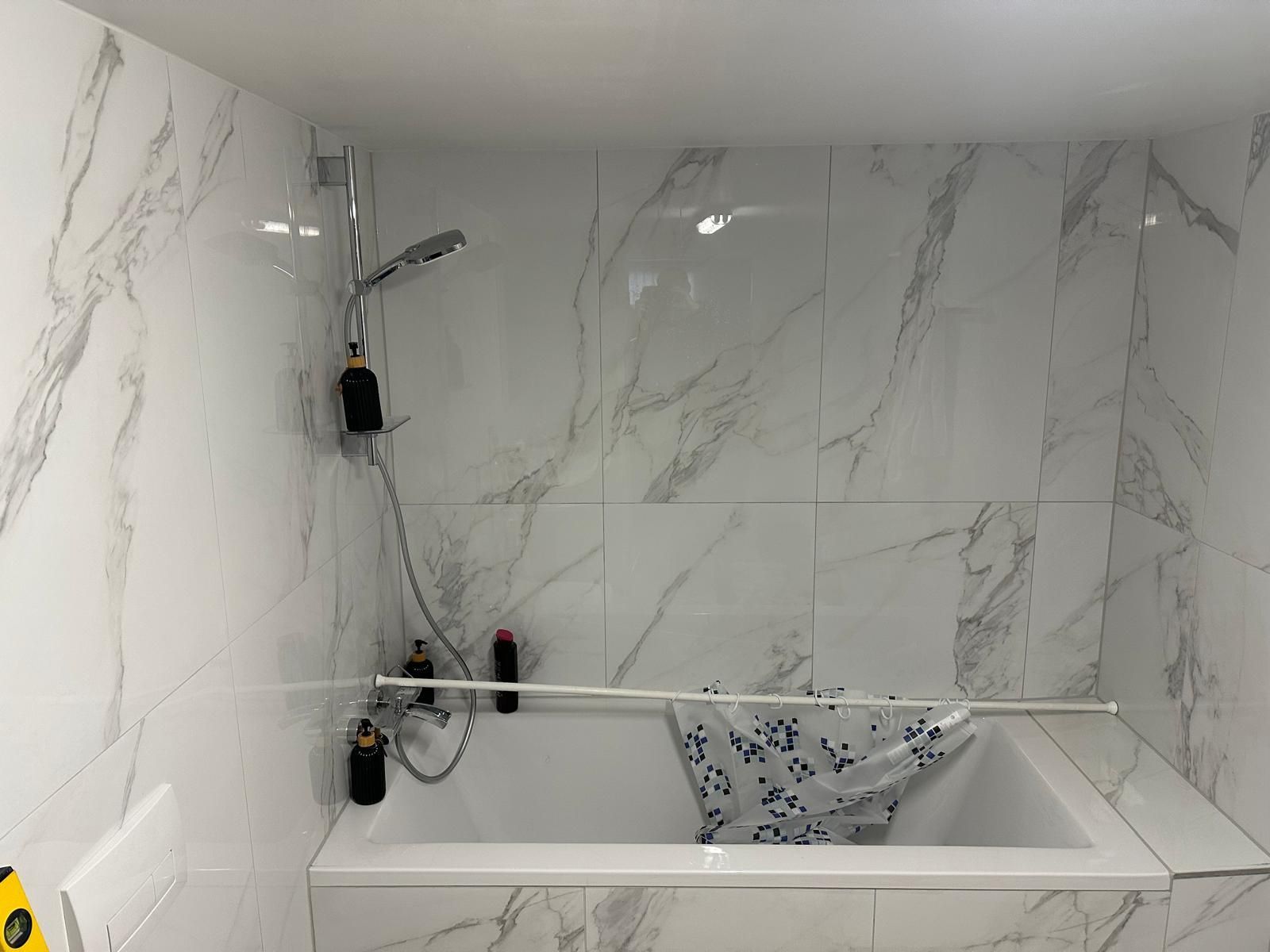 Badezimmer mit weißen Marmorfliesen, Badewanne und Dusche. Duschkopf und Pflegeprodukte hängen an der Wand.