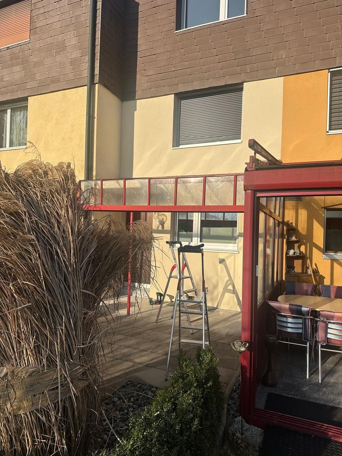 An einem Gebäude mit gelben und braunen Wänden wird an einer roten Markise mit Glaspaneelen gearbeitet.