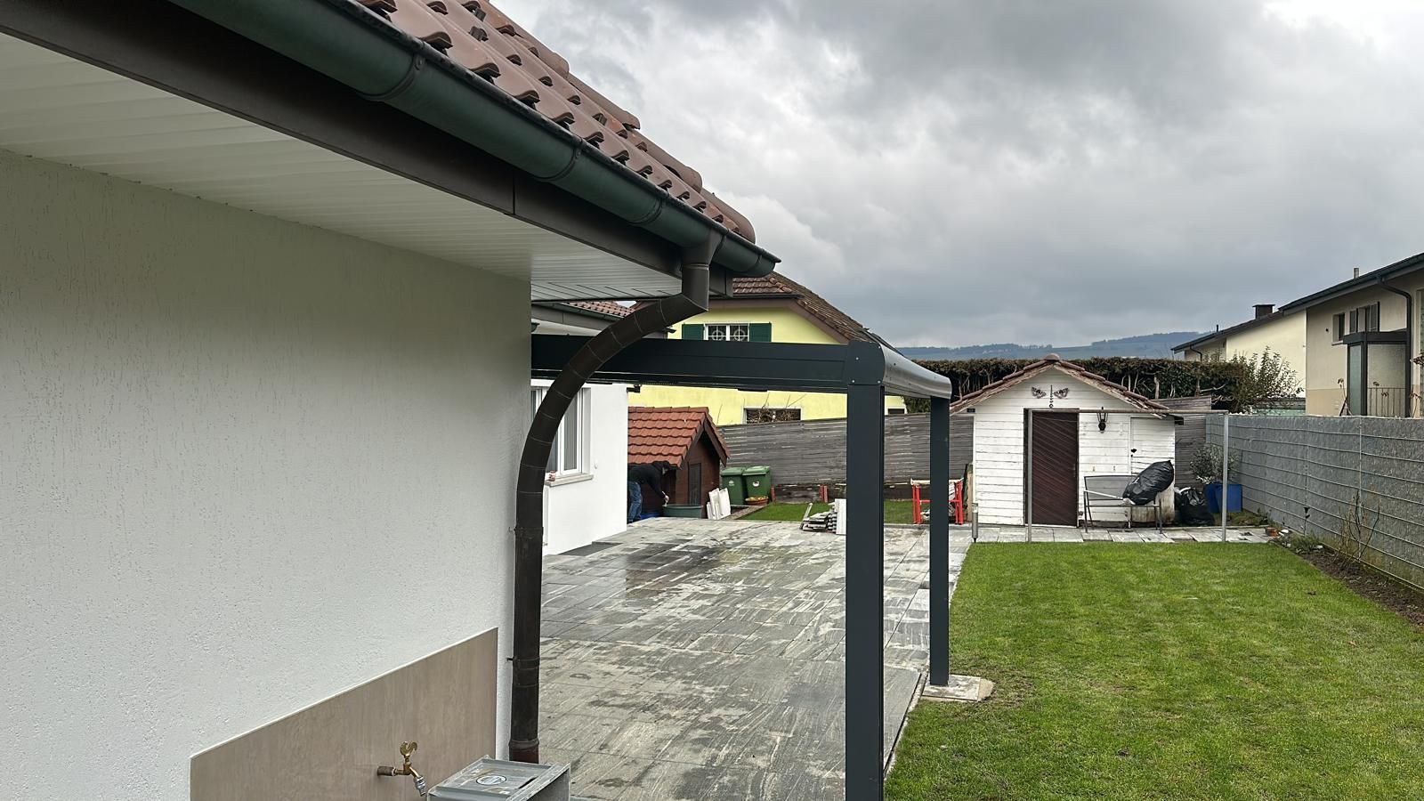 Hinterhofszene mit einem Haus, einer Terrasse, einem Rasen und einem kleinen Schuppen unter einem bewölkten Himmel.