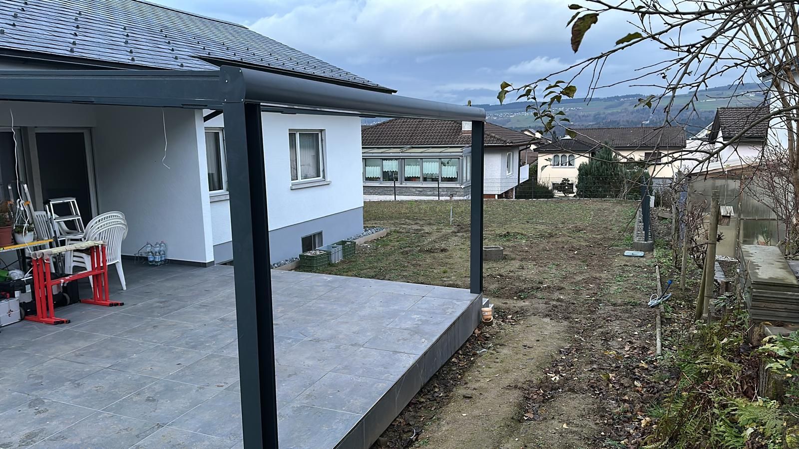 Ein graues Terrassendach bedeckt eine Betonterrasse neben einem weißen Haus. Im Hintergrund sind ein Hof mit kahlem Gras.