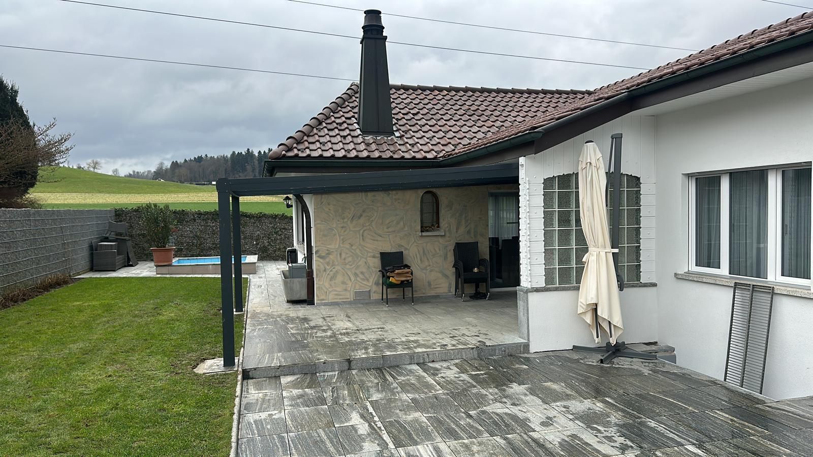 Hinterhofterrasse mit überdachtem Sitzbereich und Blick auf einen Rasen und ein entferntes grünes Feld.