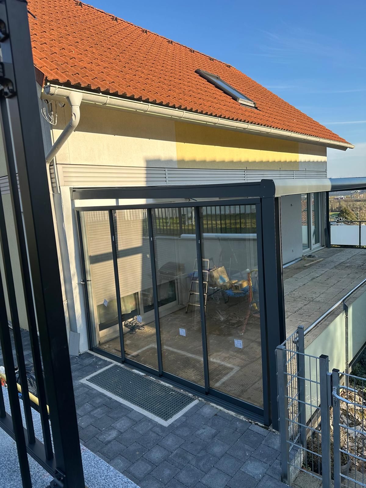 Ein moderner Wintergarten mit Glaswänden, der an ein Gebäude mit rotem Ziegeldach angebaut ist.