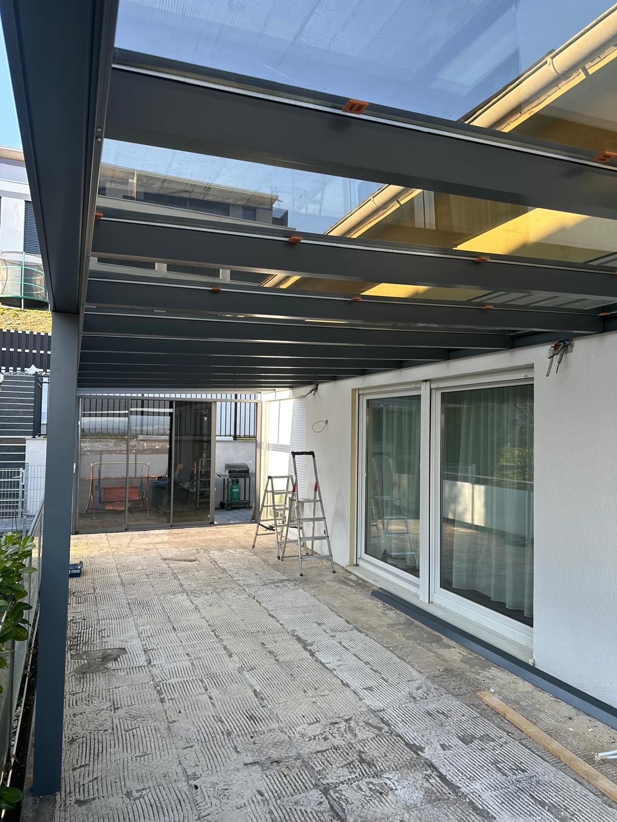 Eine Außenterrasse mit durchsichtigem Dach, grauen Stützbalken & hellem Steinboden. Es gibt Glasschiebetüren und eine Leiter.
