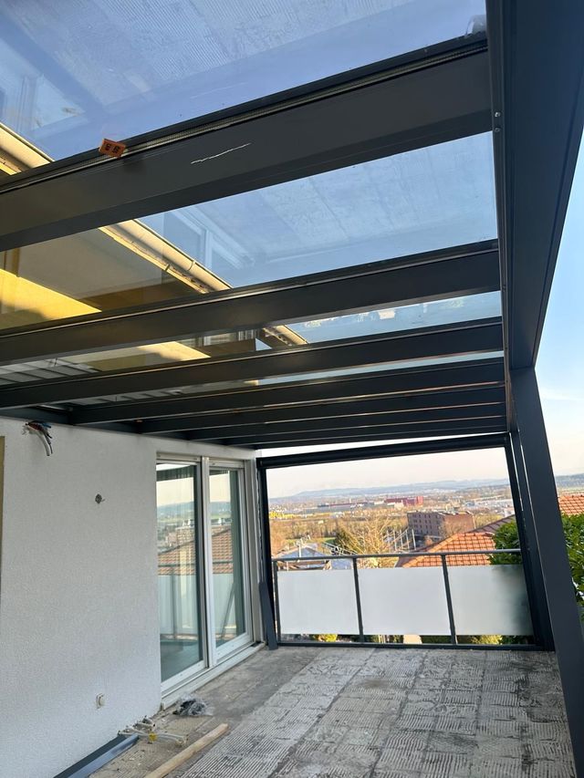 Eine moderne Terrasse mit Glasdach und Blick auf die Stadt. Ein grauer Metallrahmen stützt das Dach und umgibt die Terrasse.