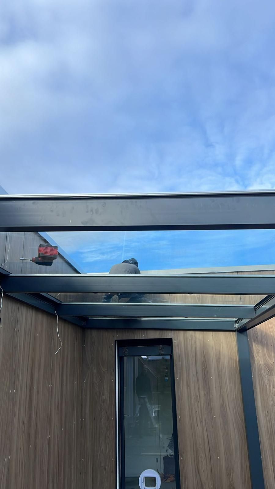 Außenkonstruktion mit Glasdach, brauner Holzwand und dunklem Rahmen vor blauem Himmel.