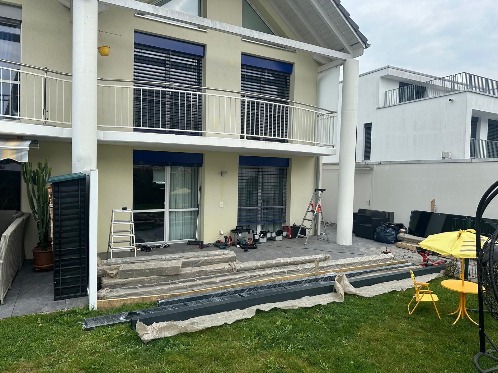 Zweistöckiges Haus mit Balkonen und blauen Fensterläden. Bauwerkzeuge stehen auf der Terrasse.