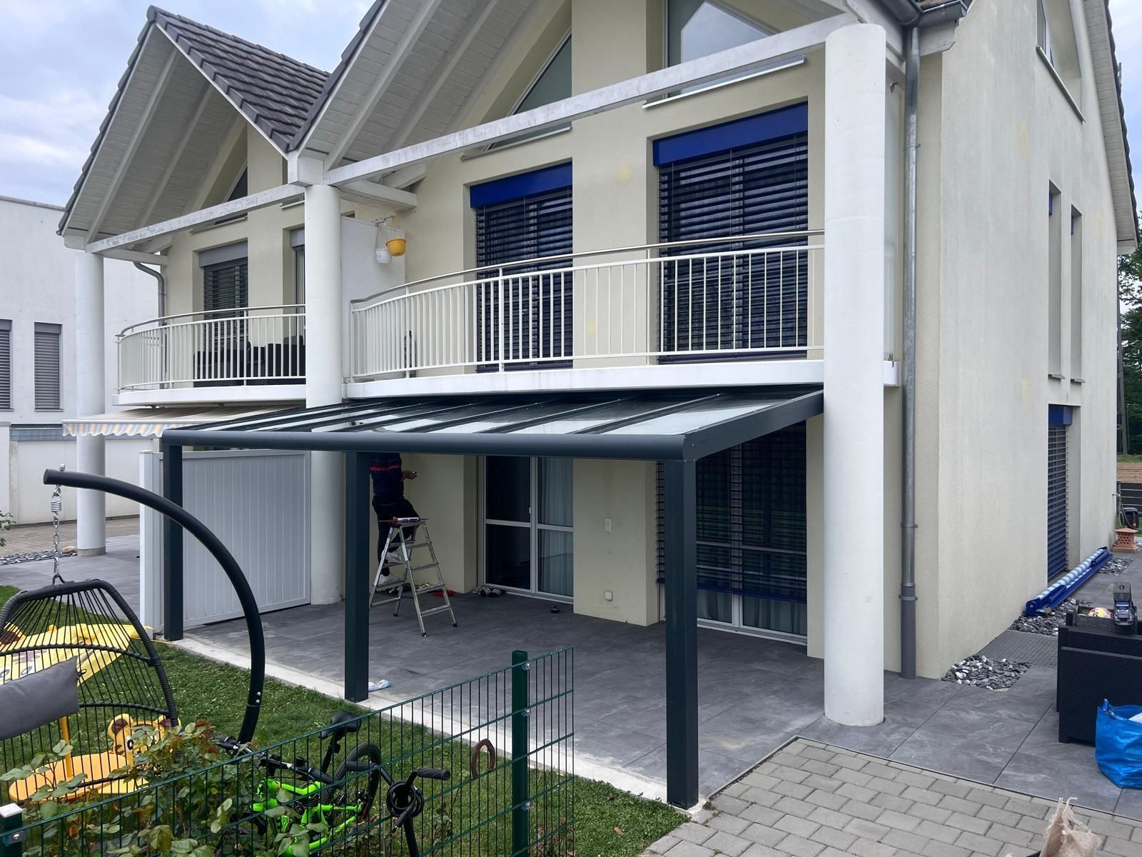 Beiges Haus mit einer Terrassenüberdachung über einer Terrasse mit Pflastersteinen. Balkone im zweiten Stock haben Geländer.