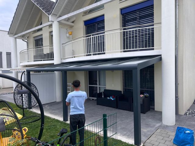 Eine Person installiert eine graue Terrassenüberdachung auf einem zweistöckigen Gebäude mit Balkon und blauen Fensterläden.