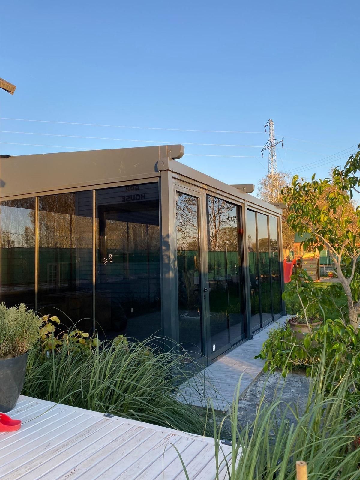 Moderner Außenraum mit Glaswänden und Dach, umgeben von Pflanzen und einer weißen Terrasse. Sonnenlicht erhellt die Szene.