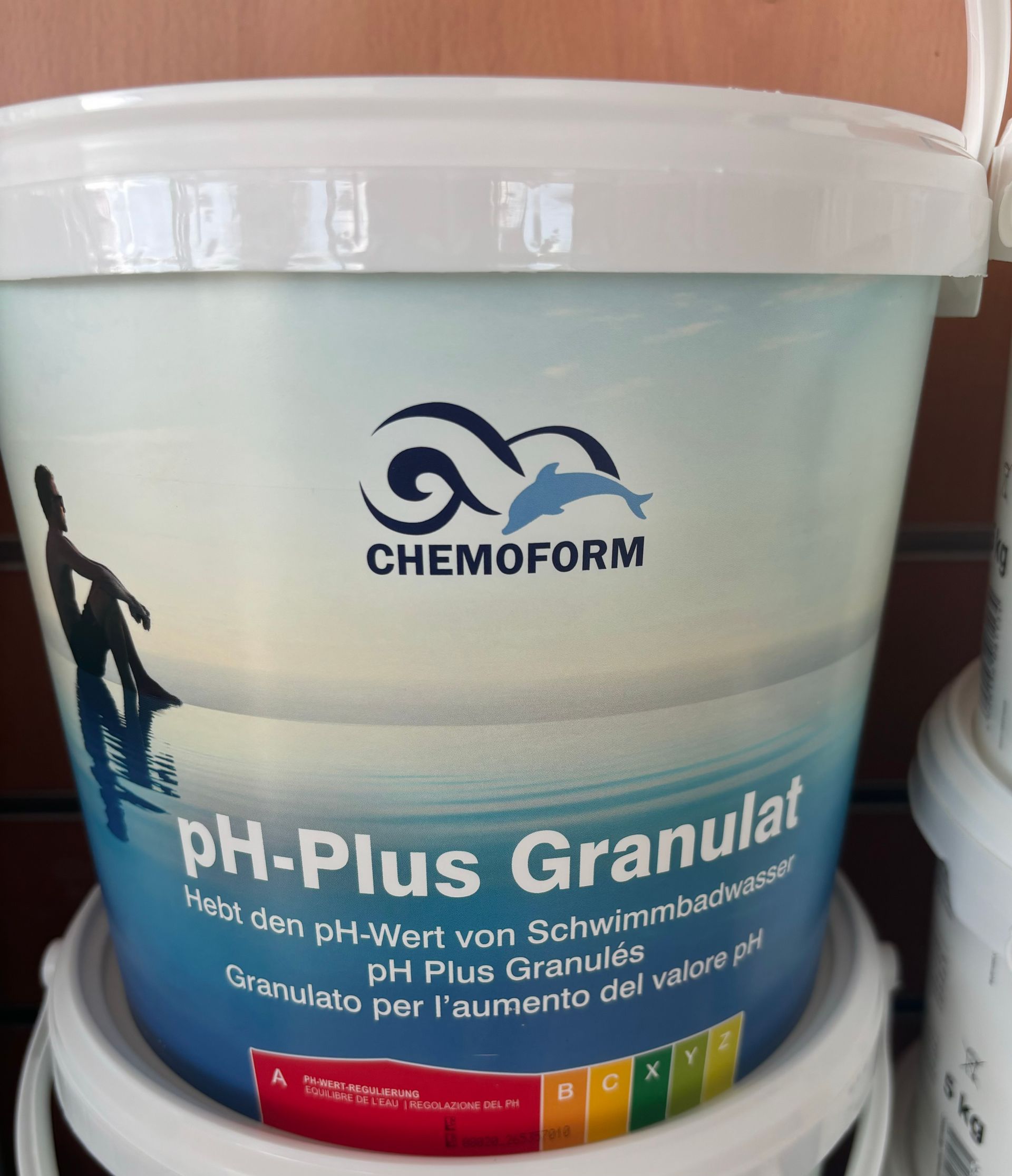 Seau blanc de produit chimique granulaire pour piscine Chemform pH-Plus avec étiquette bleue, posé sur une surface en bois.