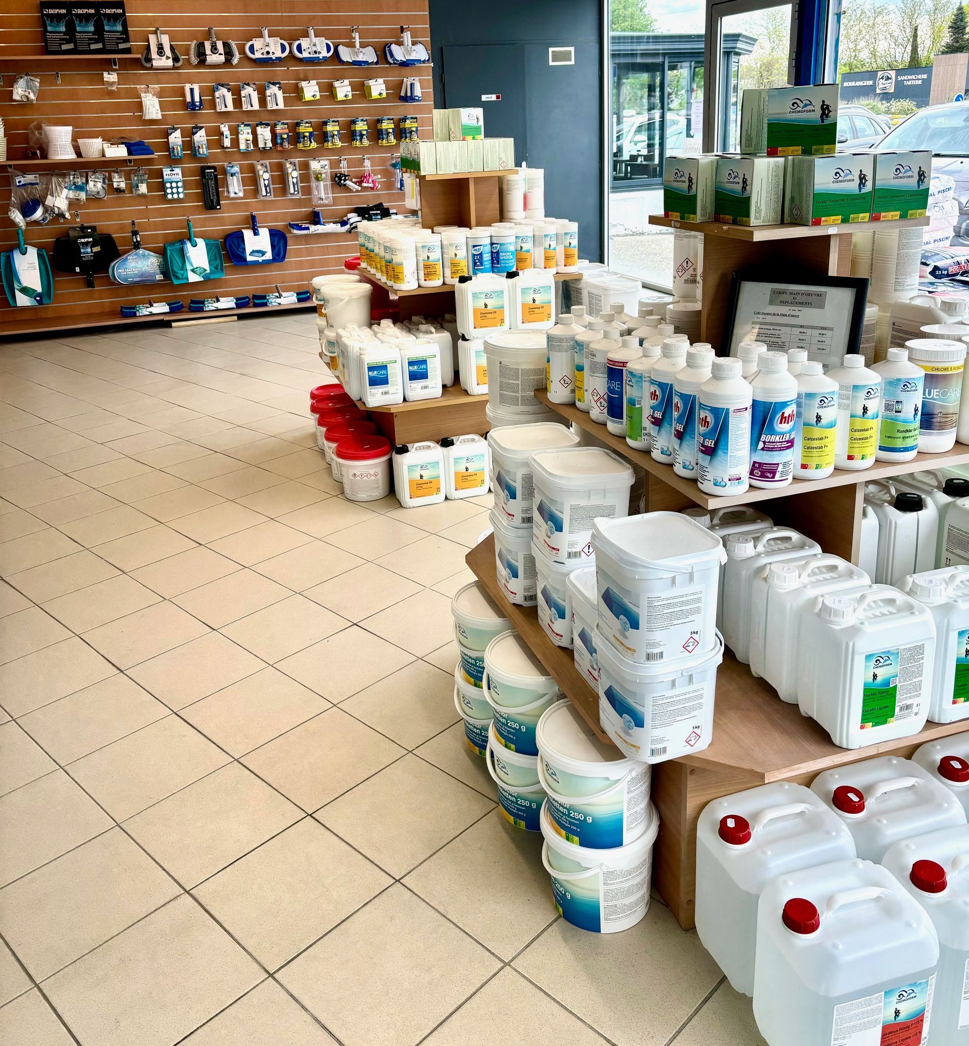 Allée d'un supermarché avec des produits de nettoyage empilés, des produits en bouteille et des étagères remplies de bocaux et d'articles ménagers.