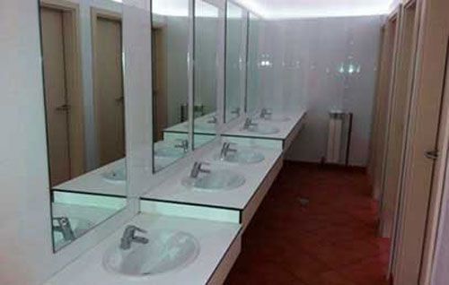 Baño público con una larga fila de lavabos, espejos y puertas de cubículos de color beige sobre un suelo de baldosas.