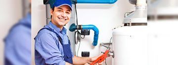 Técnico con uniforme y gorro azules inspeccionando calentador de agua blanco y tuberías azules.