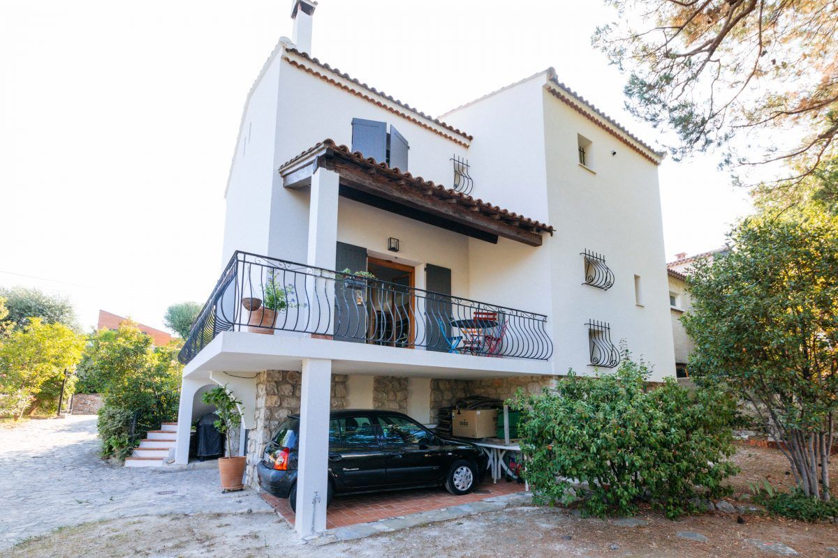 Maison sur trois niveau, une voiture est garée sous la terrasse et la terrasse est couverte, on y voit également des ouvertures avec des volets battants