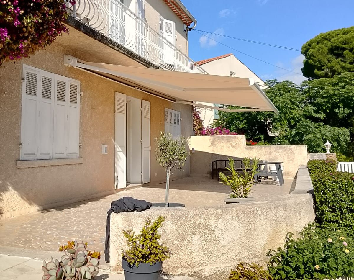 Maison beige à deux étages avec volets blancs et balcon, auvent au-dessus d'une terrasse avec table et chaises, ciel bleu.