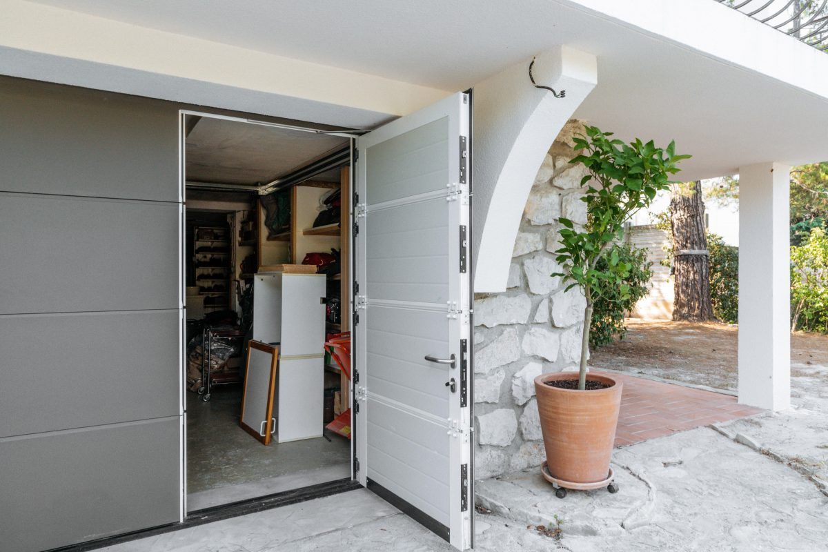 Porte de garage avec une porte à ouverture individuelle ouverte. La maison a un mur en pierre et on y voit une grande plante.
