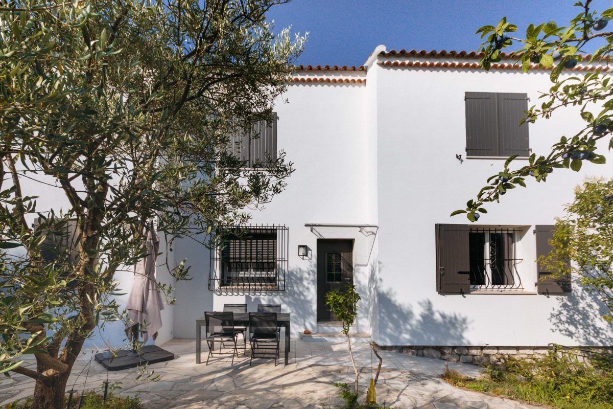 Maison blanche avec quatre fenêtres et une porte d'entrée, trois des fenêtres sont dotées de volets battants. Un table et des chaises sont disposées sur une terrasse et un arbre est présent au premier plan.