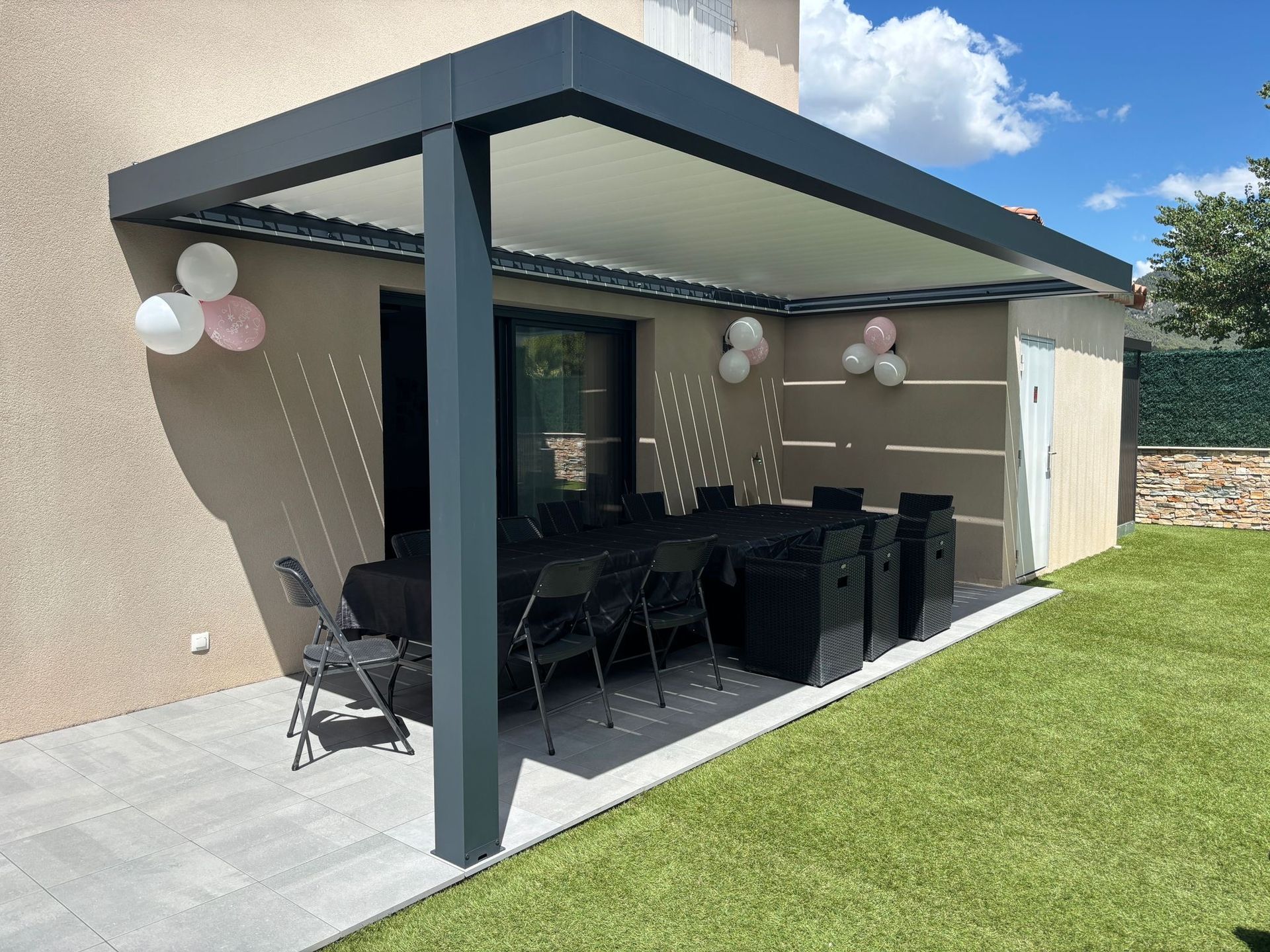 Terrasse avec table et chaises sous une pergola, décorée de ballons. Table et chaises noires sur une terrasse pavée.