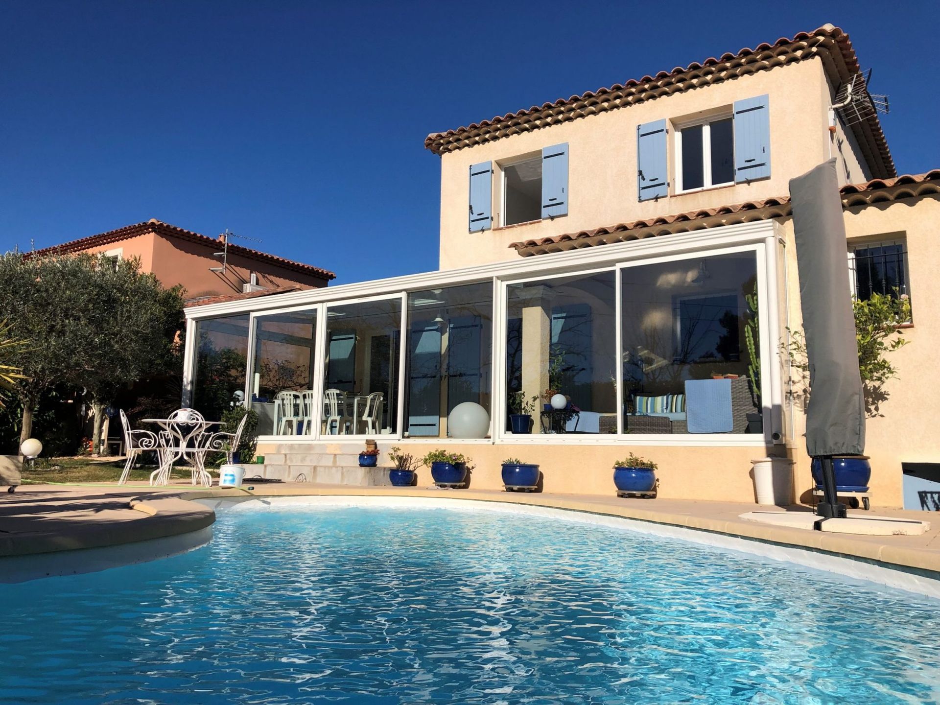 L'image représente une piscine située devant une maison avec une grande véranda. Elle met en valeur les éléments extérieurs, la piscine reflétant le ciel.