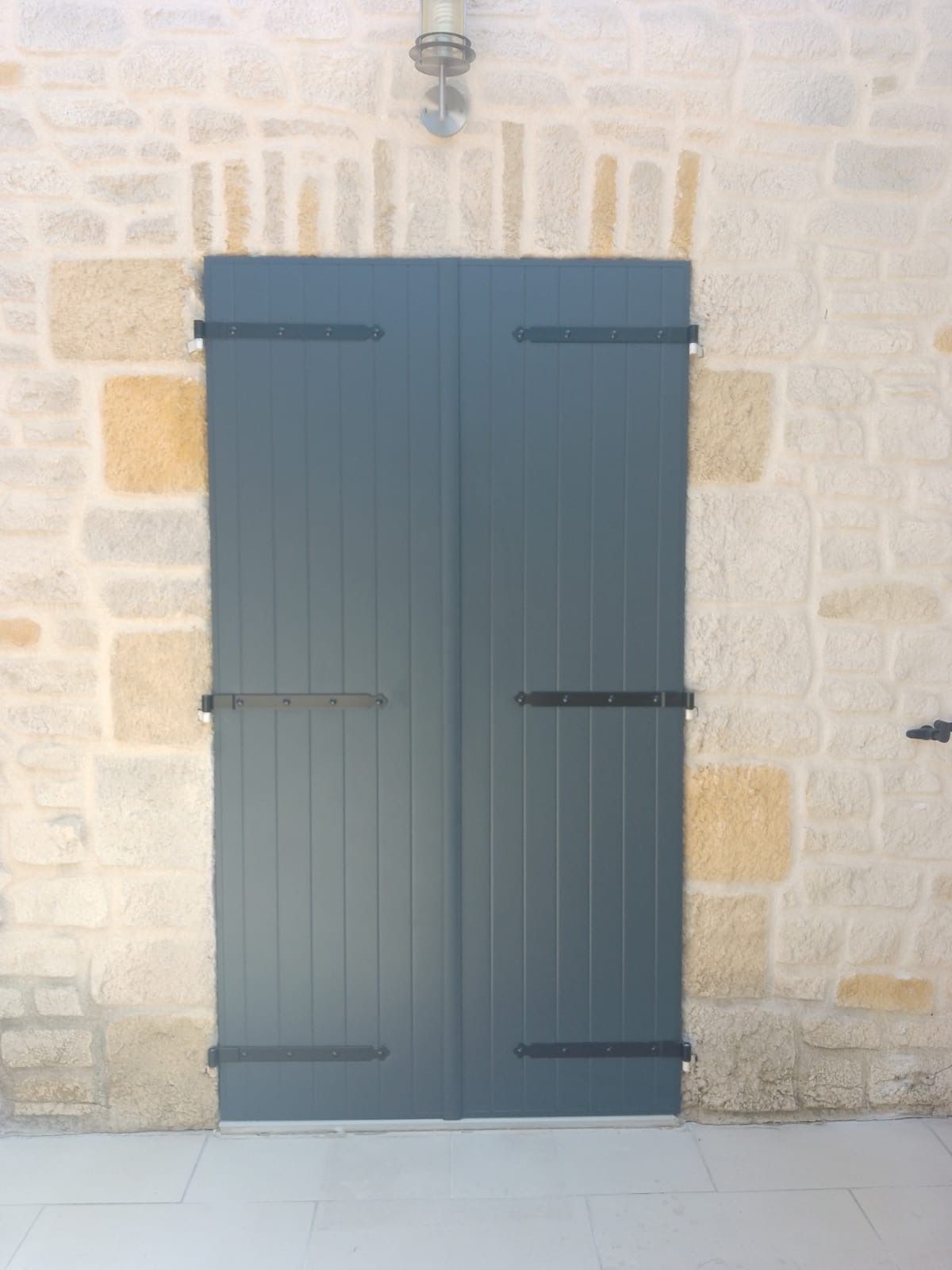 Deux portes à volets bleu-gris avec quincaillerie noire, encastrées dans un mur de pierre.