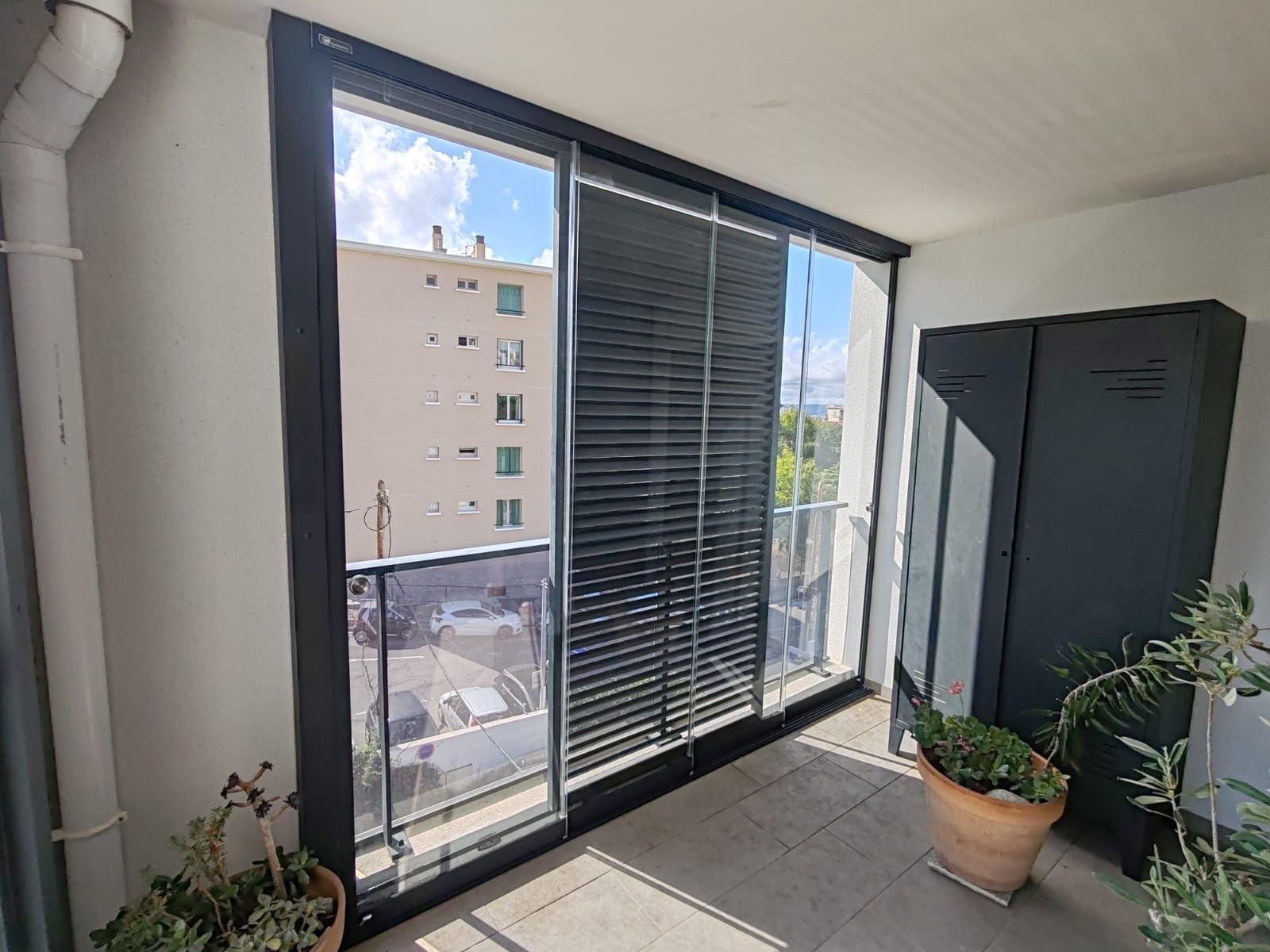Balcon avec grandes portes coulissantes en verre et stores partiels ; vue sur les immeubles et une plante en pot.
