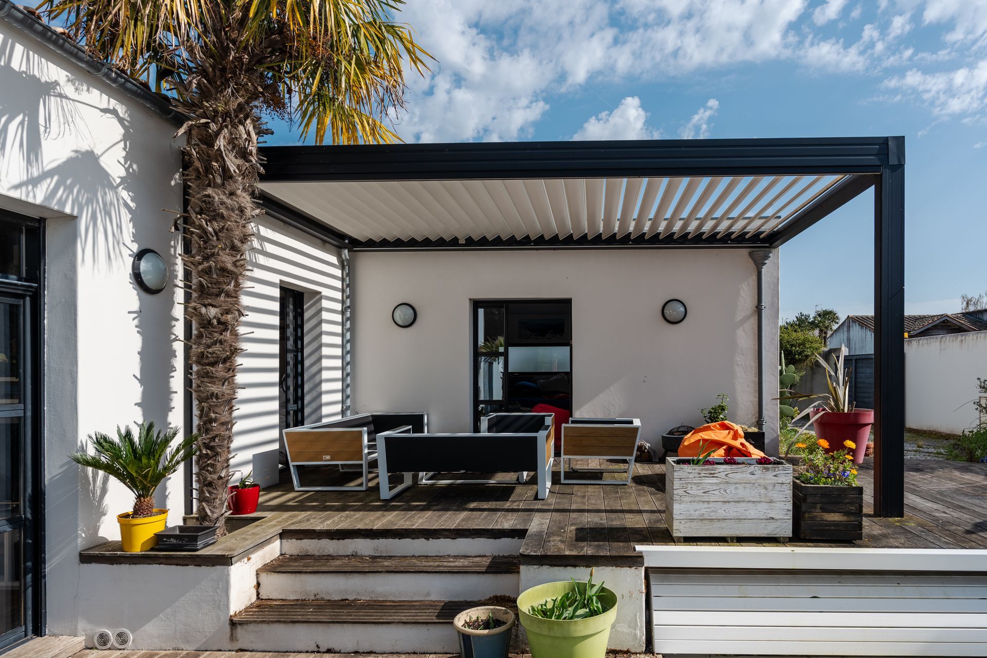 Une maison blanche dotée d'une terrasse équipée de chaises. La propriété est située en plein air, entourée de plantes et sous un ciel dégagé. Elle présente des éléments architecturaux typiques des maisons résidentielles, notamment une pergola bioclimatique.