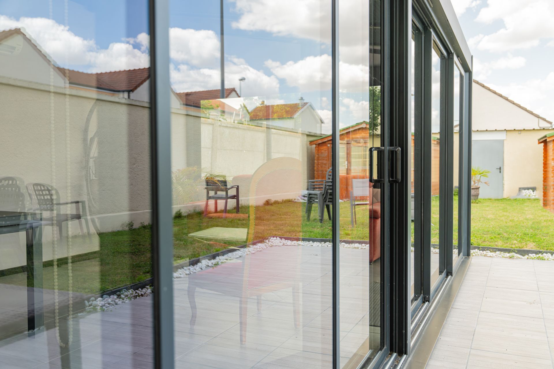 Des portes coulissantes en verre reflètent une scène de jardin avec pelouse, chaises et clôture blanche.