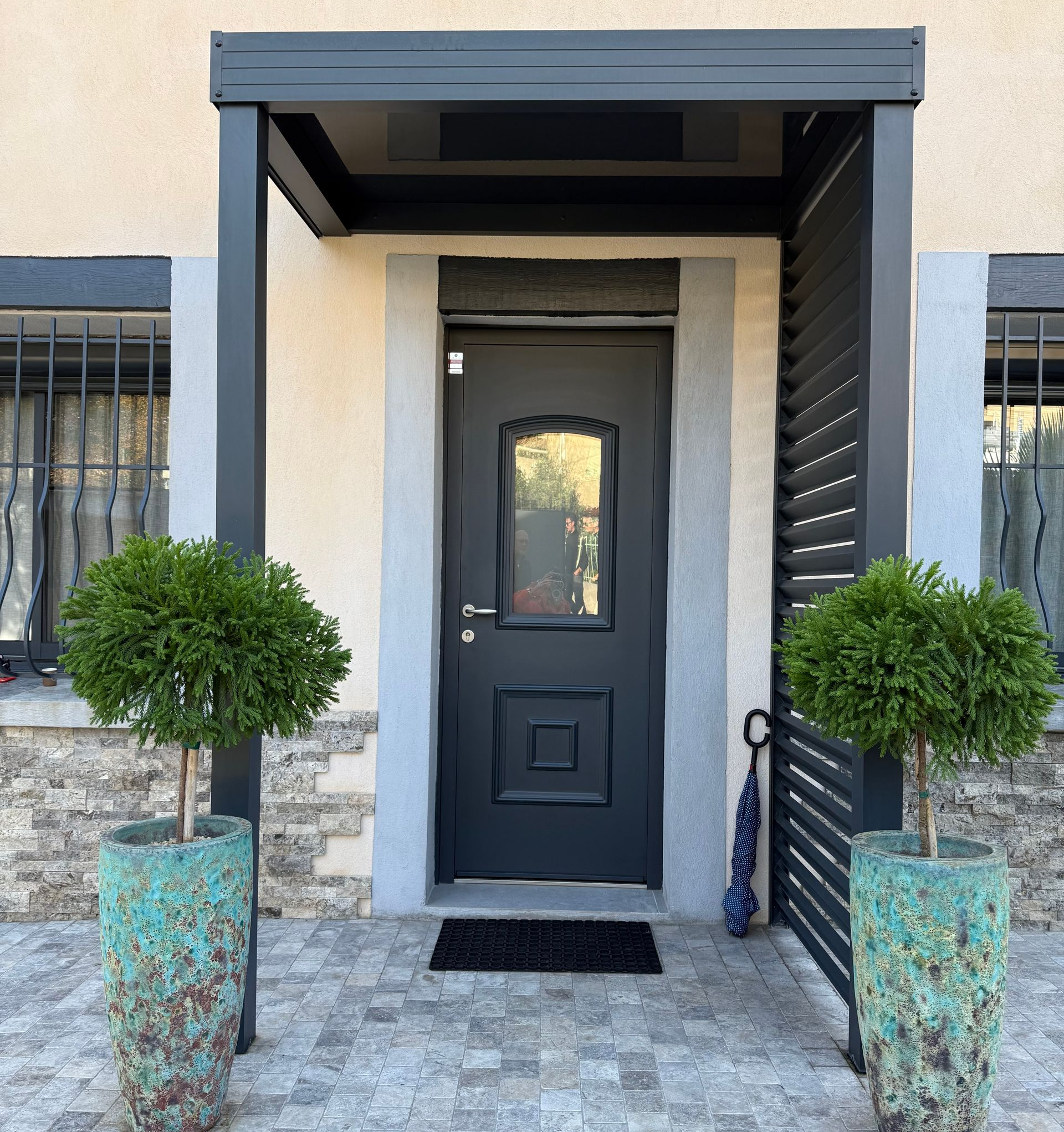 Porte grise à panneau de verre, sous un auvent sombre, flanquée d'arbres topiaires dans des pots turquoise.