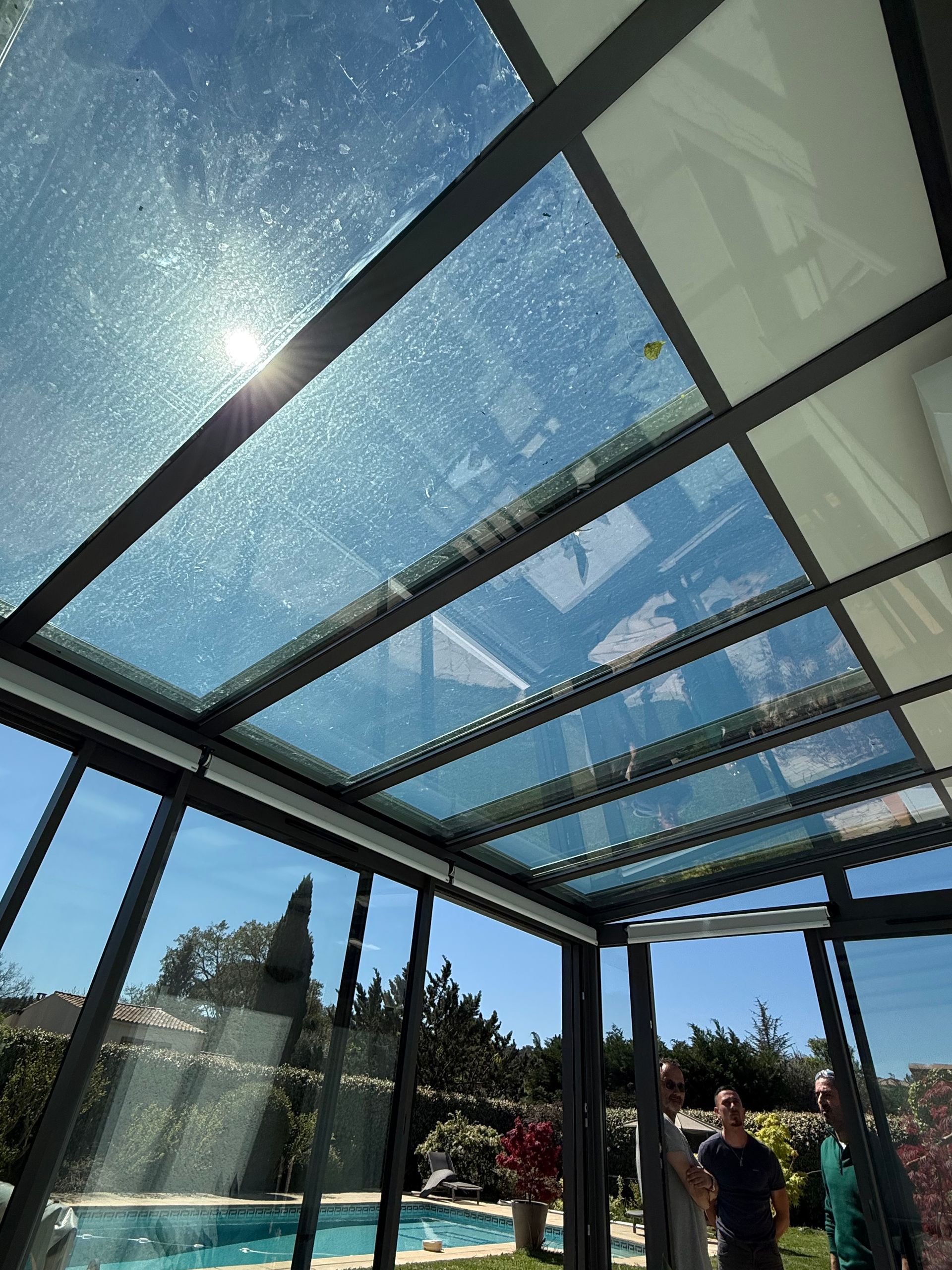 Véranda vitrée donnant sur un ciel bleu. Des personnes se tiennent à l'intérieur, contemplant la piscine et le jardin.