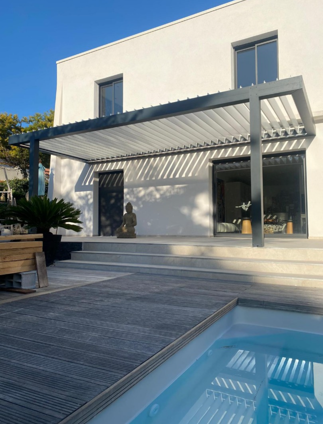 Maison blanche moderne avec une pergola surplombant un patio. Une petite piscine et une statue se trouvent devant.