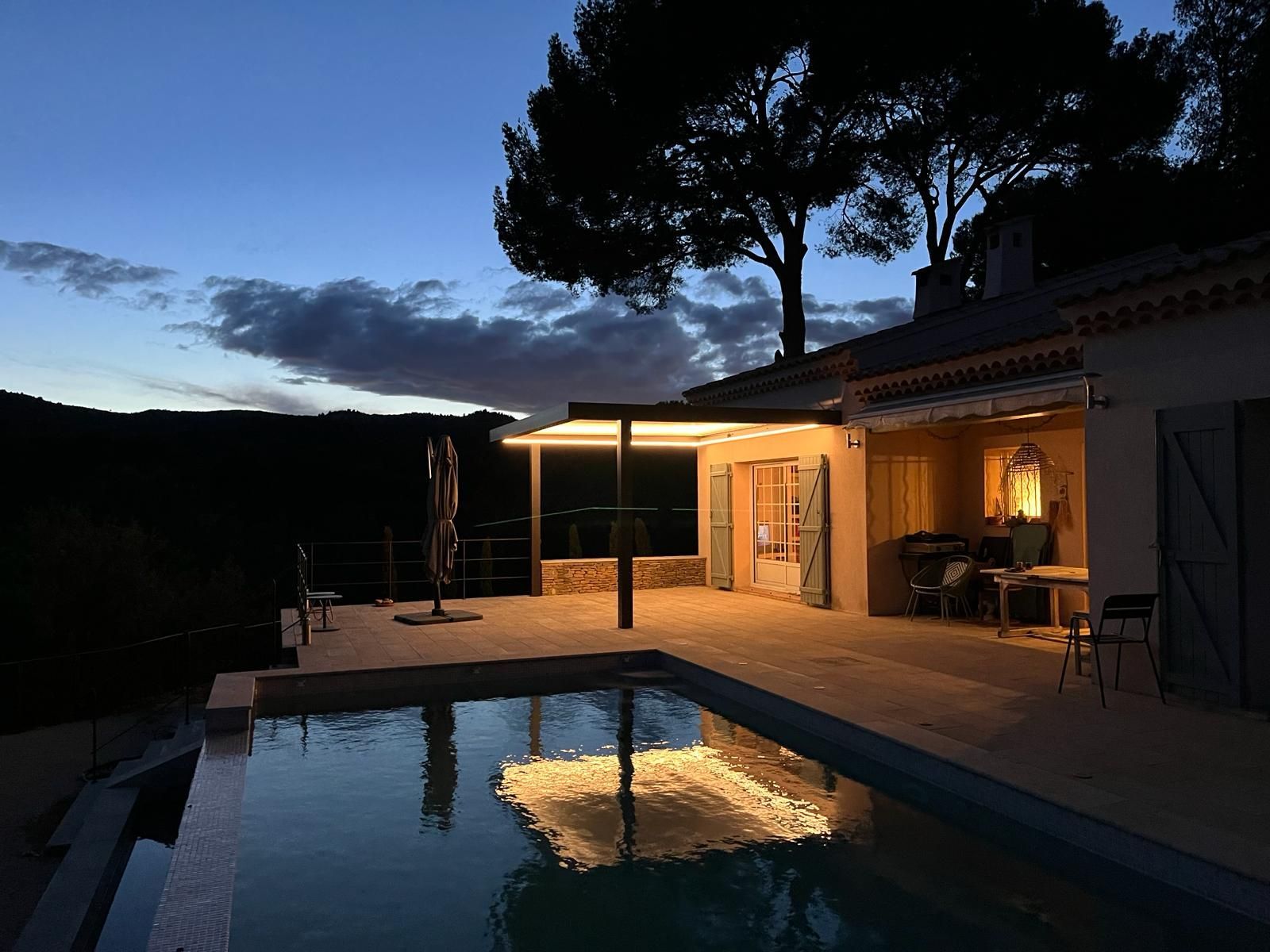 Une maison avec et pergola et une piscine la nuit. Le ciel est dégagé et la propriété est entourée d'arbres, contribuant à son ambiance sereine. Le décor évoque une atmosphère de détente, rappelant l'atmosphère d'une station balnéaire ou d'une station thermale.