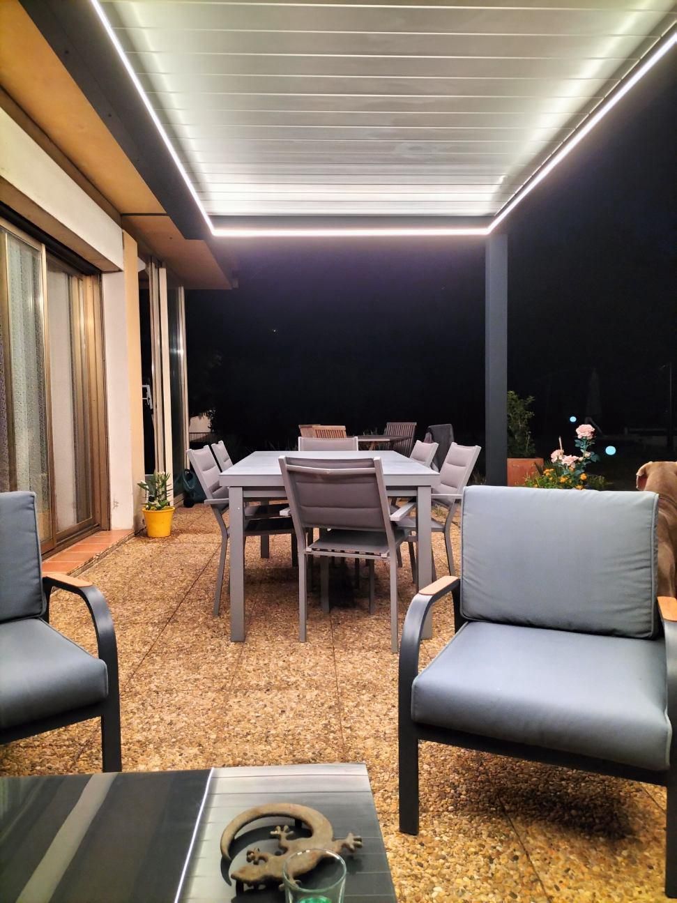 Intérieur d'une pergola avec une table et des chaises disposées sur une terrasse, suggérant un espace repas ou de détente en plein air. La scène est probablement conçue pour le confort et le style, mettant en valeur les éléments de décoration intérieure.
