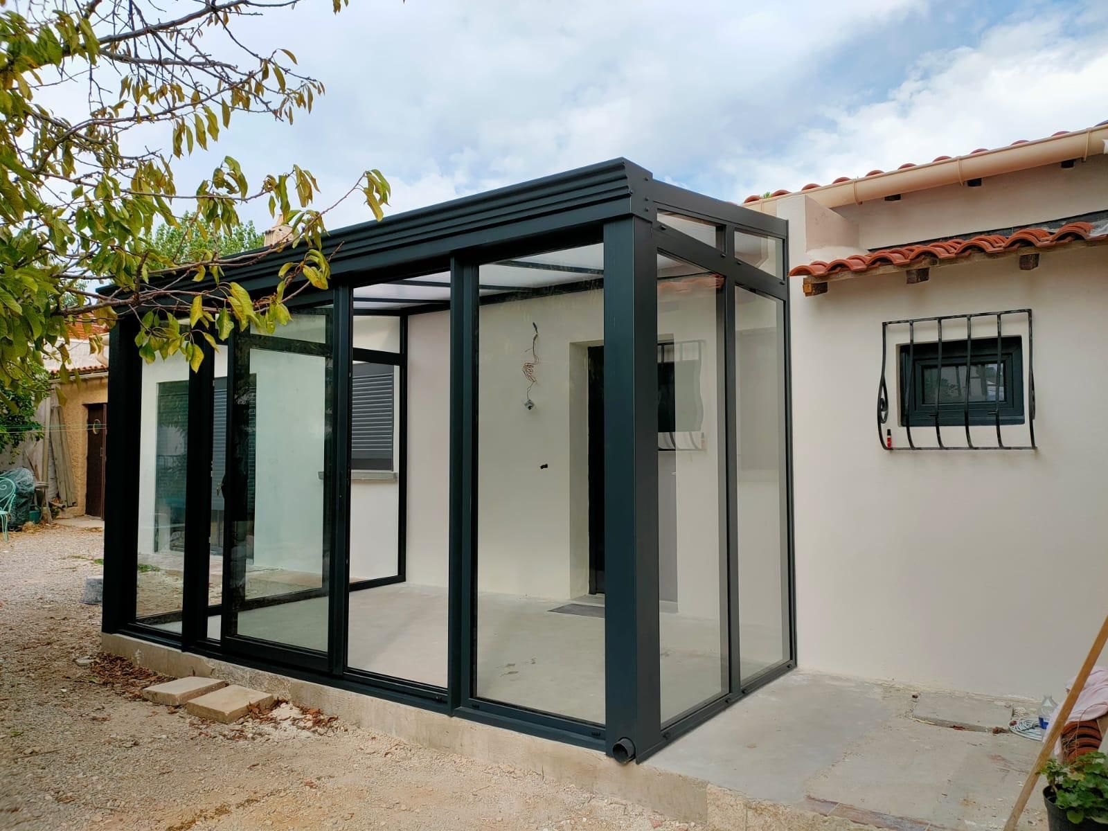 Véranda en aluminium gris devant un façade de maison avec un fenêtre protégée par des barreaux.