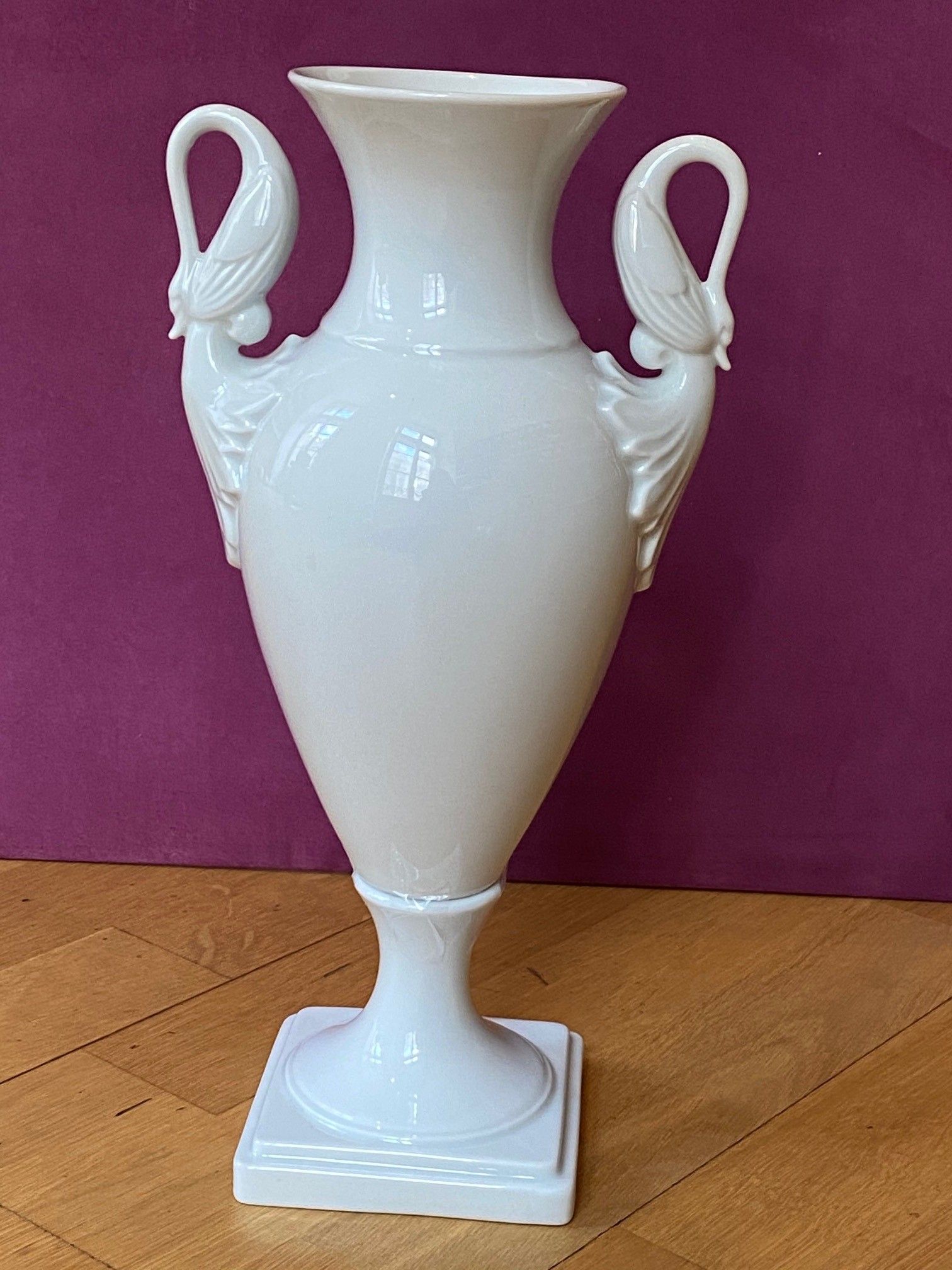 Weiße Porzellanvase mit Schwanenhenkeln auf quadratischem Sockel vor einer bordeauxroten Wand und einem Holzboden.