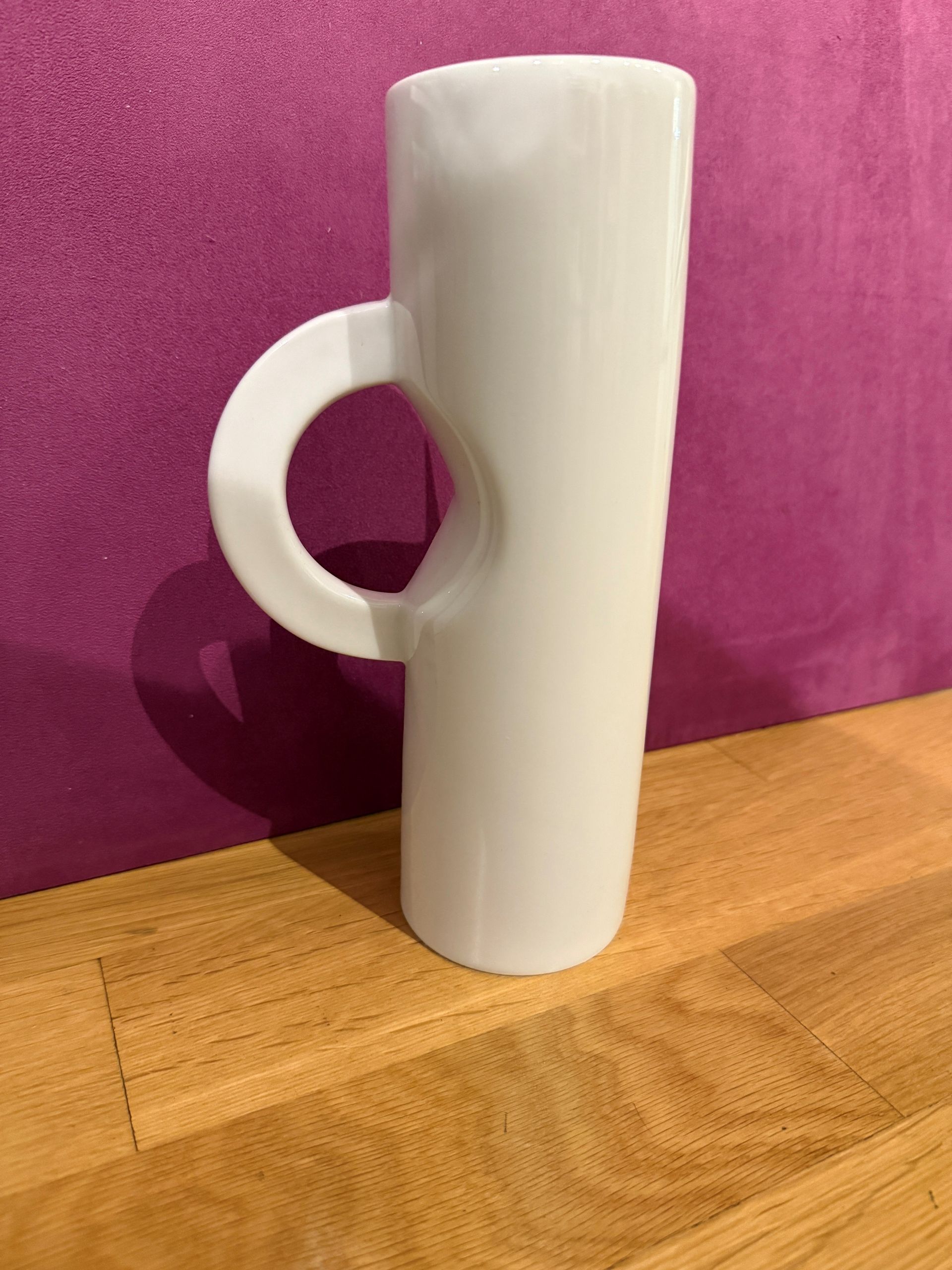 Weißer Keramikbecher mit rundem Henkel auf Holzoberfläche, violetter Hintergrund.