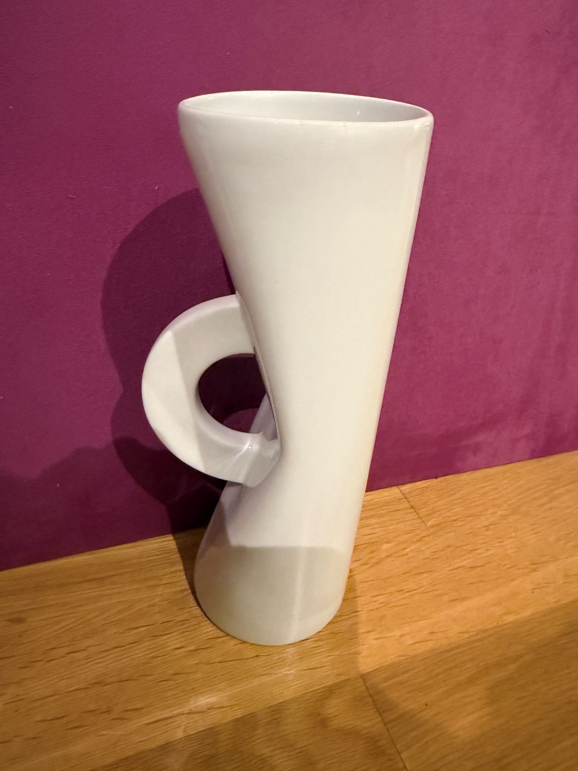 Weißer Keramikbecher mit gebogenem Henkel, der auf einer Holzoberfläche vor einer violetten Wand steht.