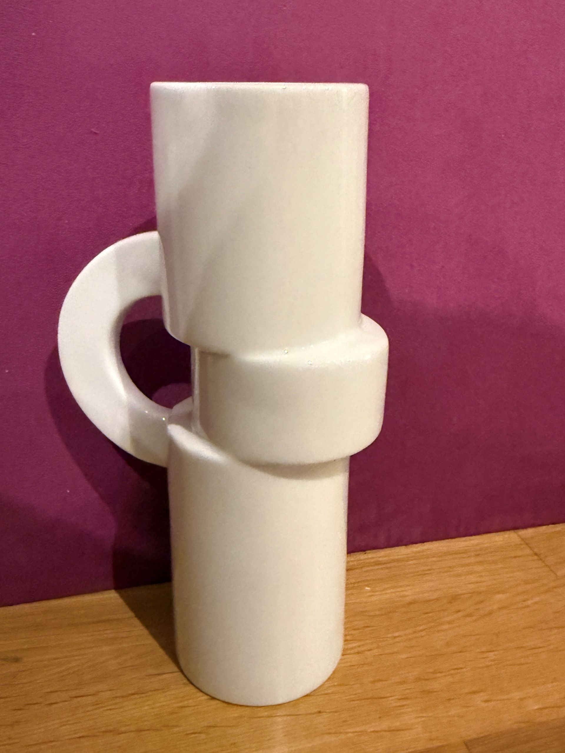 Abstrakte weiße Keramikvase mit Henkel auf Holzoberfläche, violetter Hintergrund.