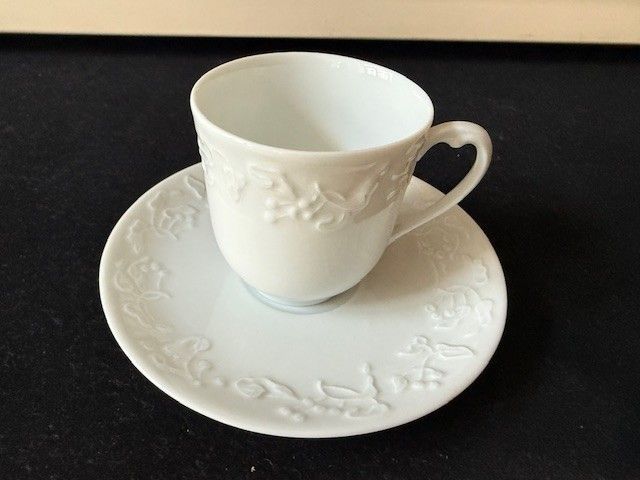 Weiße Teetasse mit Untertasse und geprägtem Blumendekor auf dunklem Untergrund.