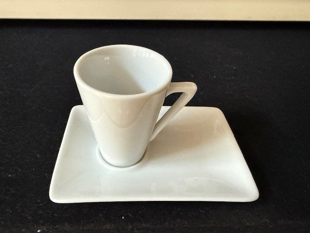 Weiße Kaffeetasse auf einer quadratischen weißen Untertasse auf schwarzem Untergrund.