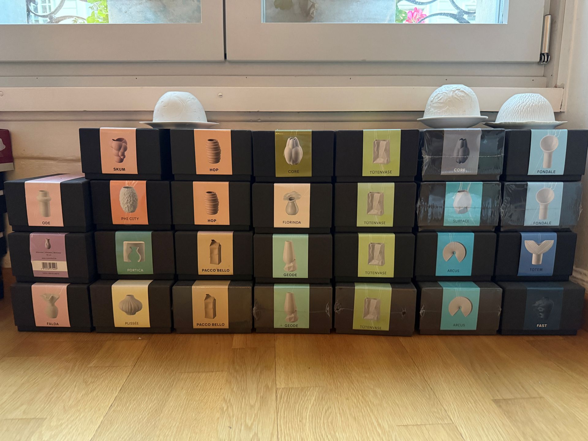 Reihen schwarzer Schachteln mit bunten Etiketten, gekrönt von weißen Zierkugeln, angeordnet vor einem Fenster.