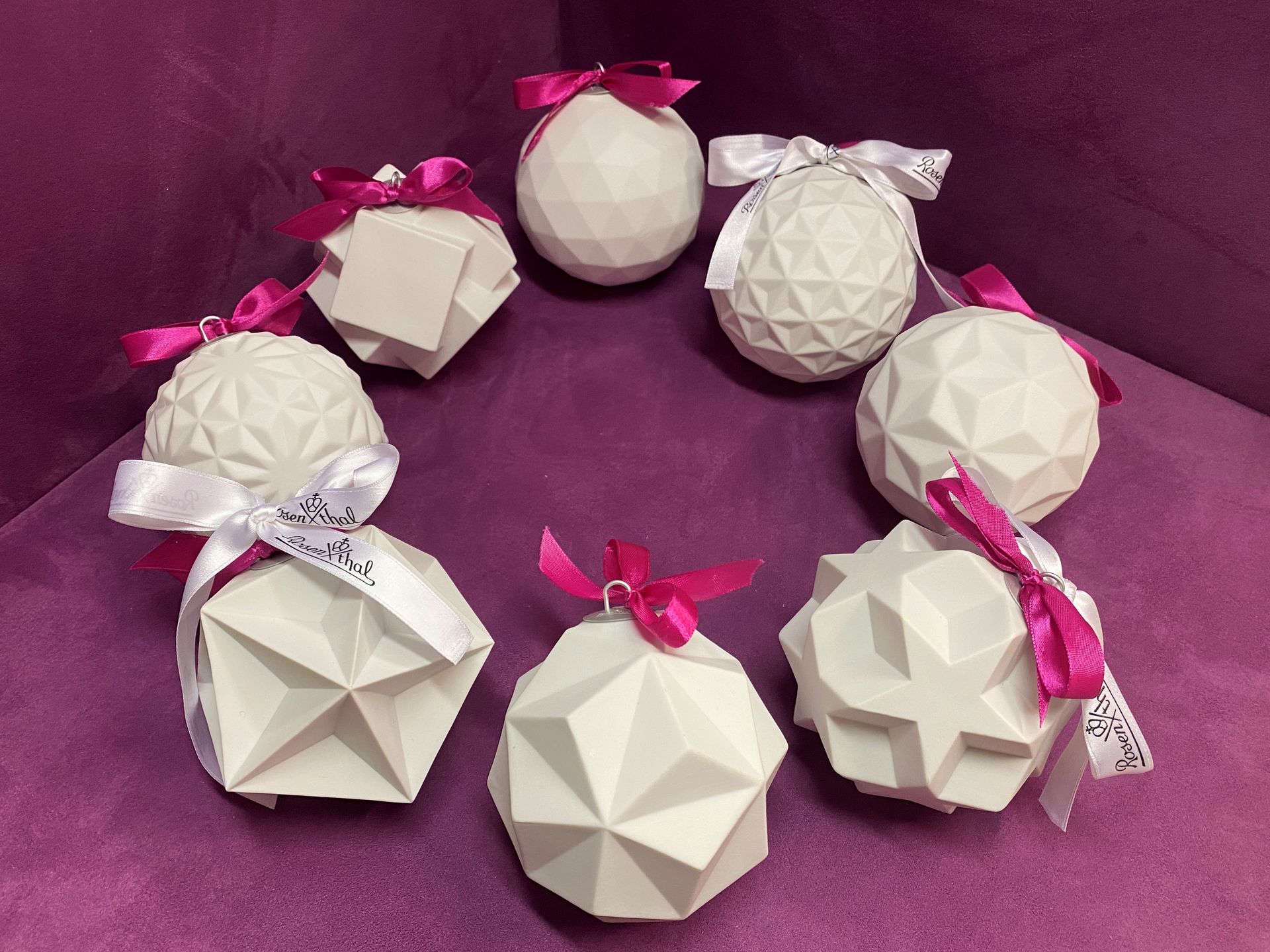 Weiße geometrische Ornamente mit rosa oder weißen Bändern, die auf einem violetten Untergrund kreisförmig angeordnet sind.