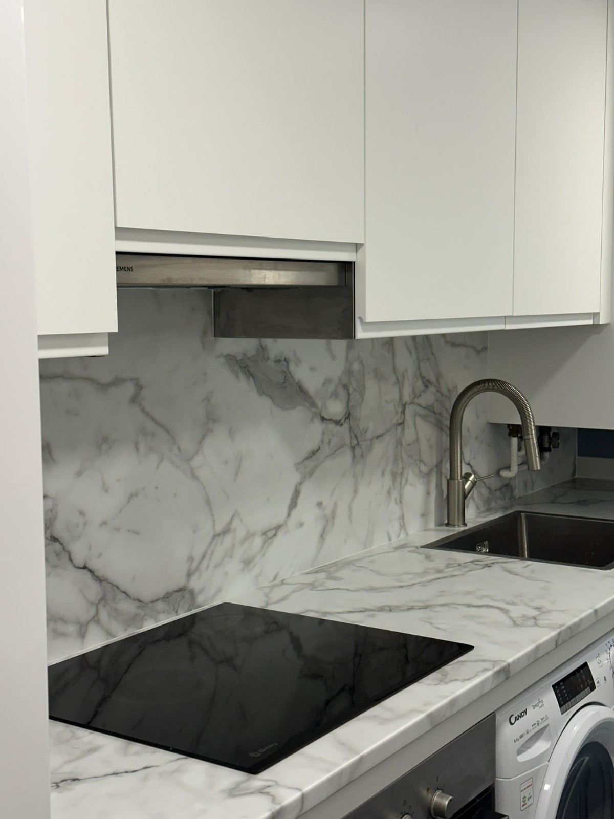 Cocina moderna con armarios blancos, salpicadero de mármol, placa de cocina negra y fregadero de acero inoxidable bajo una campana extractora
