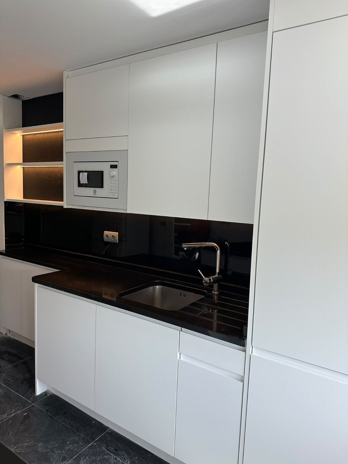 Cocina moderna con armarios blancos, encimeras negras, microondas empotrado y fregadero de acero inoxidable