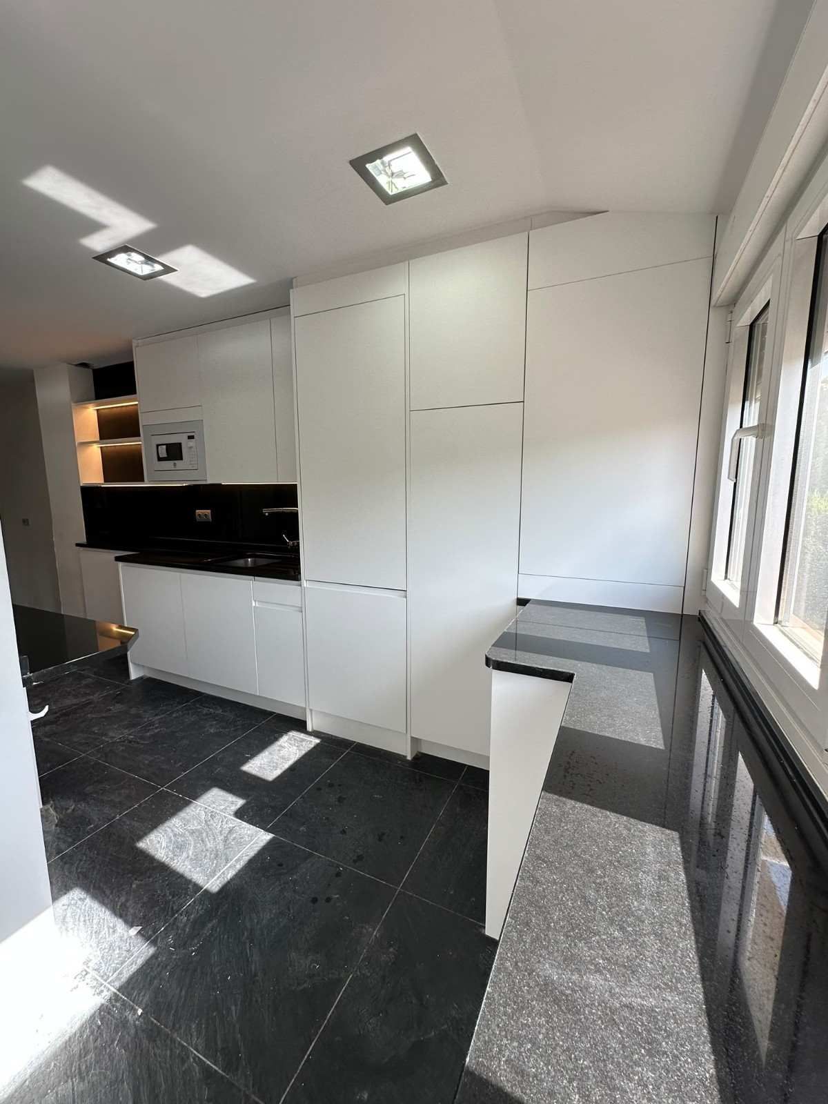 Cocina moderna blanca con suelo de baldosas negras, armarios empotrados y grandes ventanales a lo largo de la pared derecha