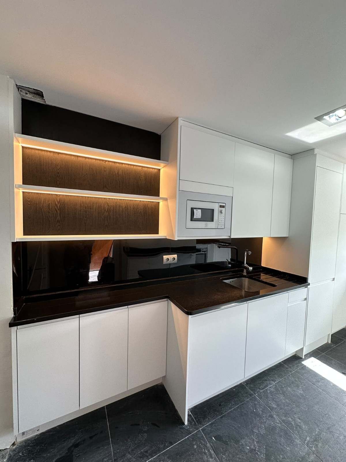 Cocina moderna con armarios blancos, encimeras negras, estantes abiertos con iluminación y microondas empotrado.