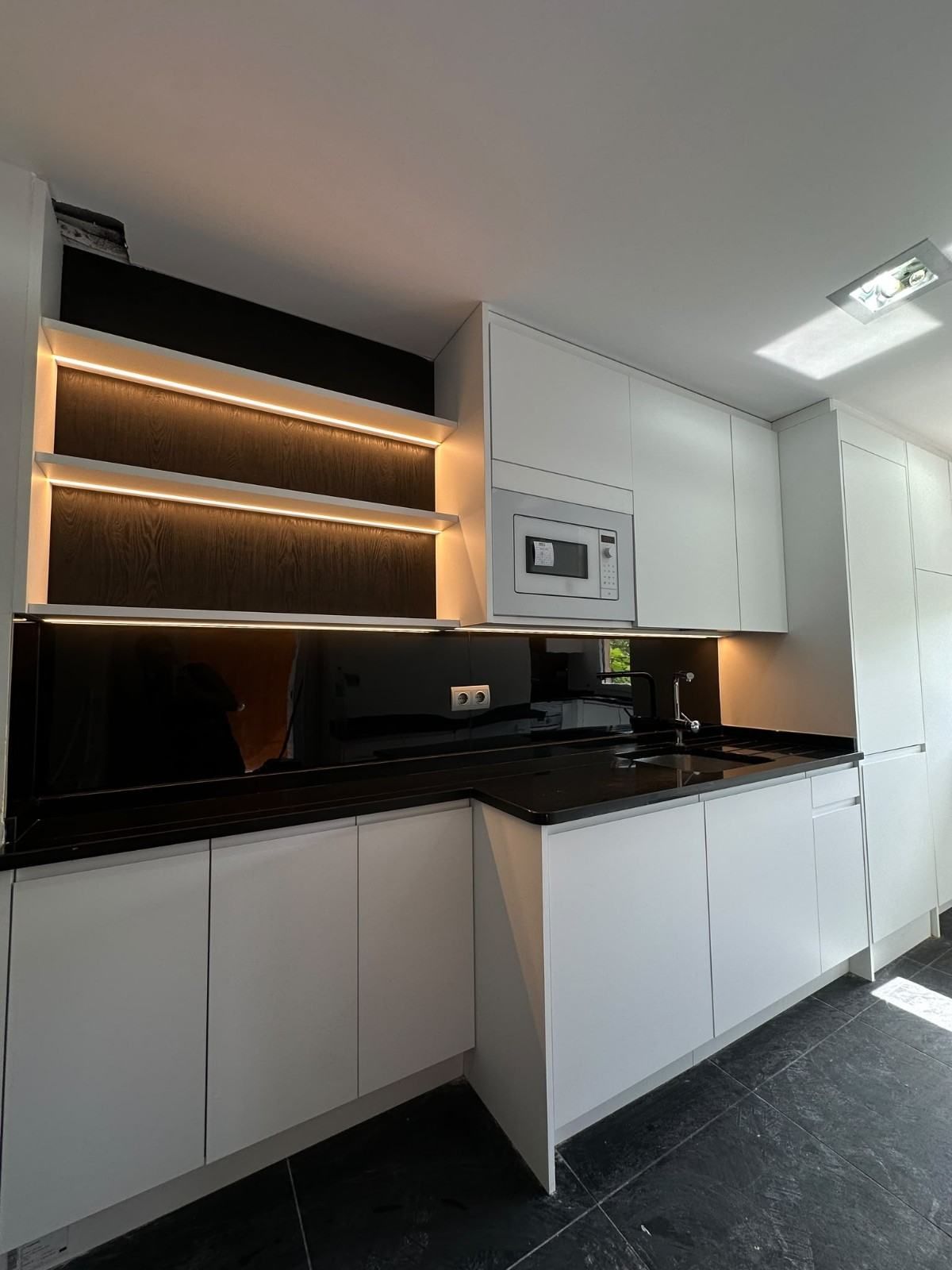 Cocina moderna con armarios blancos, encimera negra, estantes abiertos iluminados y horno y microondas empotrados.