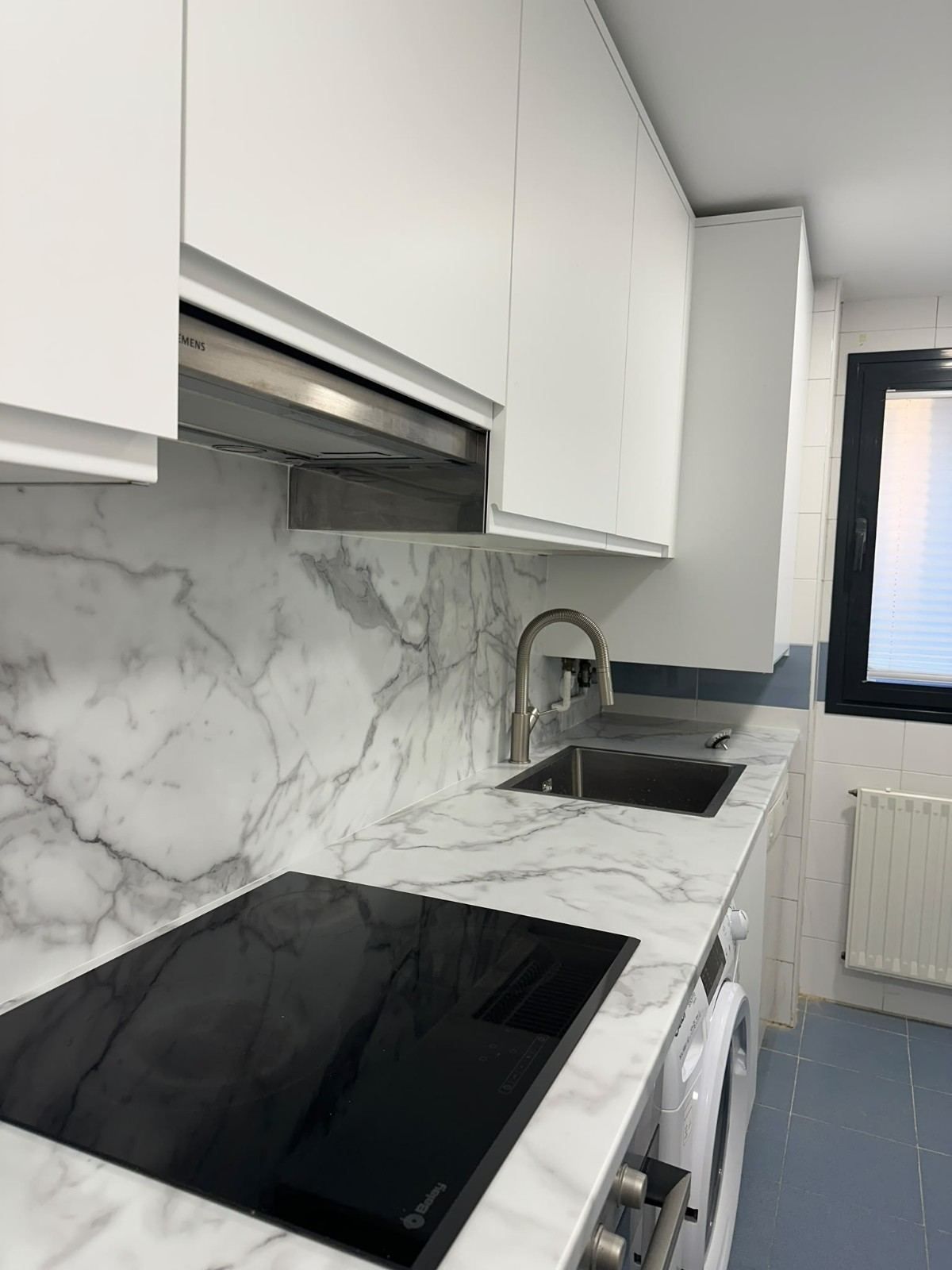 Cocina moderna con armarios blancos, salpicadero de mármol, placa de cocina negra, fregadero y baldosas azules en el suelo.